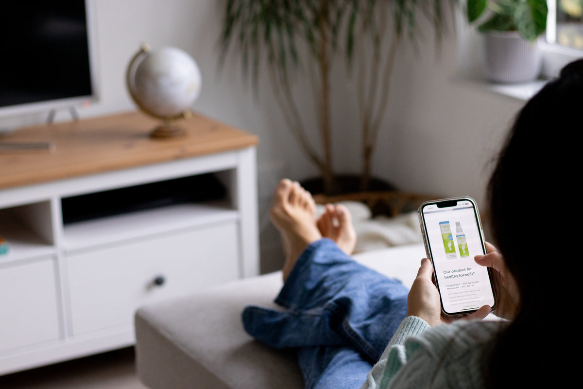 Iemand bekijkt FunghiClear® voetverzorgingsproducten op een smartphone terwijl hij thuis uitrust.